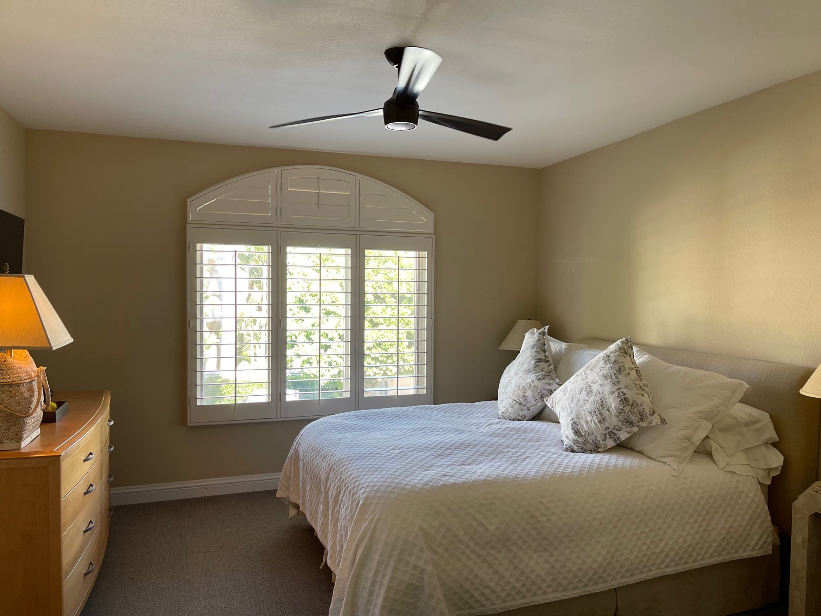Three-blade ceiling fan with a light installed over the bed in a bedroom with an arched shuttered window