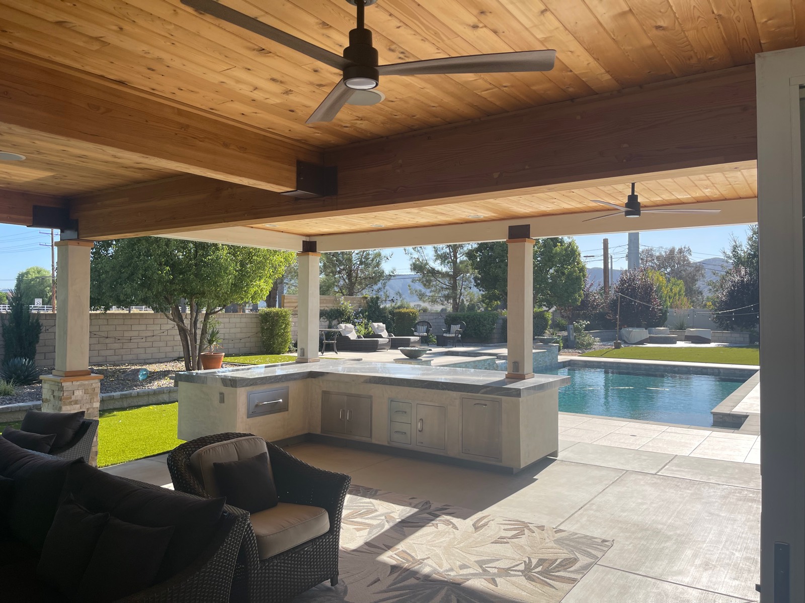 Two ceiling fans installed under a wood-beamed patio cover beside an outdoor kitchen and a pool