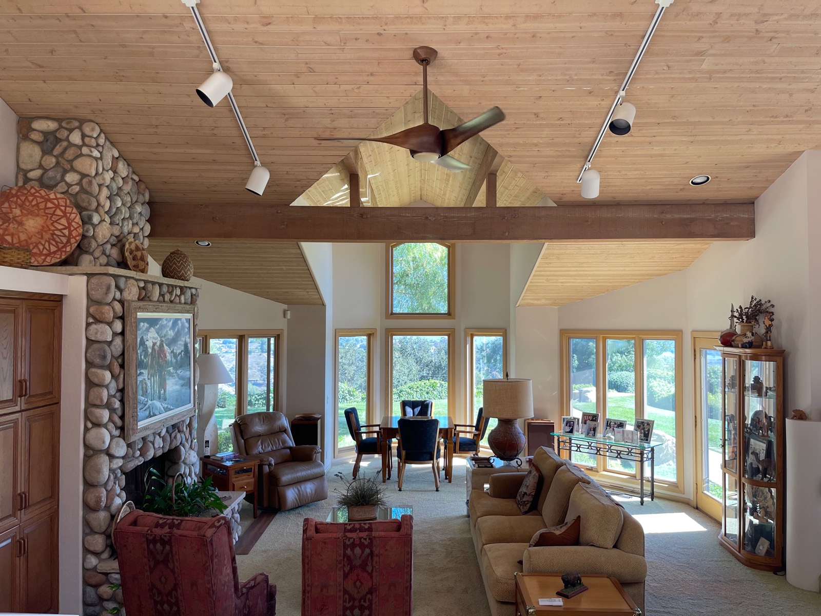 Wood-bladed ceiling fan hung from a sloped wood ceiling in a great room with a river-rock fireplace and track lighting